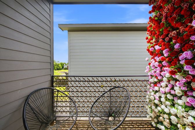 Small suburban balcony with two black circular wire chairs, lattice railing and a vibrant vertical rose wall of red, pink and white blooms against beige siding and blue sky.