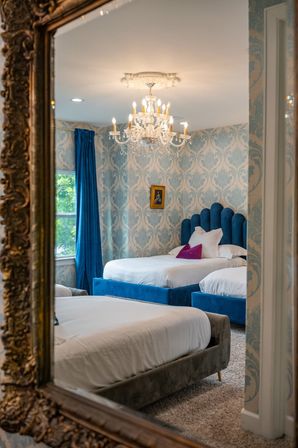 Reflection of a boutique-style bedroom interior with blue velvet beds, damask wallpaper, crystal chandelier and ornate gilded mirror.