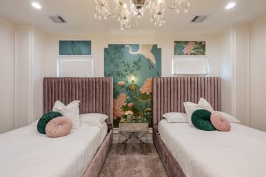 Modern luxury twin bedroom interior with plush mauve velvet headboards, green and blush round pillows, mirrored nightstand and brass wall lamp centered on a teal botanical mural with crane, crystal chandelier overhead