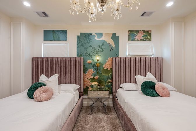Modern luxury twin bedroom interior with plush mauve velvet headboards, green and blush round pillows, mirrored nightstand and brass wall lamp centered on a teal botanical mural with crane, crystal chandelier overhead