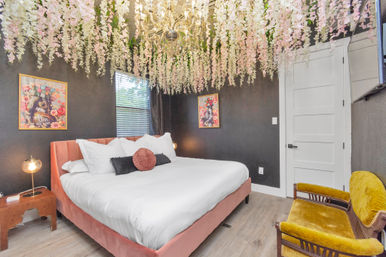 Boutique bedroom with hanging pink and white wisteria canopy and crystal chandelier, blush velvet king bed with white linens and decorative pillows, dark textured accent wall, vintage mustard velvet loveseat and framed floral art.