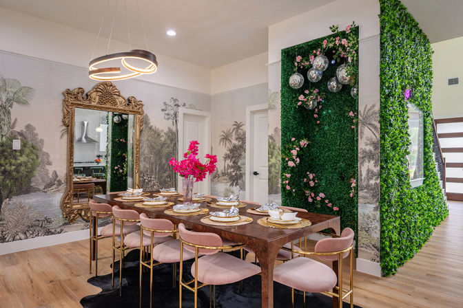 Luxurious dining room with long wooden table set for eight, blush pink velvet chairs with gold frames, ornate gold mirror, modern ring chandelier, vertical green wall covered in foliage and pink roses with hanging disco balls, tropical wallpaper and hardwood floors.