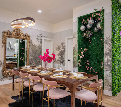 Luxurious dining room with long wooden table set for eight, blush pink velvet chairs with gold frames, ornate gold mirror, modern ring chandelier, vertical green wall covered in foliage and pink roses with hanging disco balls, tropical wallpaper and hardwood floors.