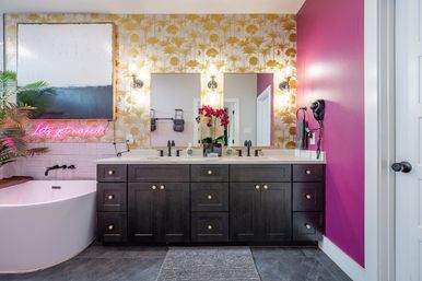 Stylish tropical-inspired bathroom with dark double-sink vanity, gold palm wallpaper, magenta accent wall, freestanding tub and playful neon "Let's get naked" sign