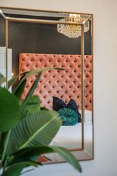 Mirror reflection of a modern bedroom featuring a tufted blush velvet headboard, crystal chandelier, teal round throw pillow on a white bed, and a lush green indoor plant in the foreground.