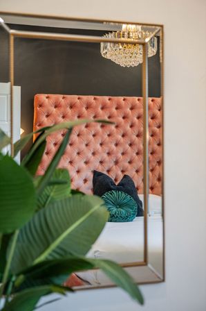Mirror reflection of a modern bedroom featuring a tufted blush velvet headboard, crystal chandelier, teal round throw pillow on a white bed, and a lush green indoor plant in the foreground.