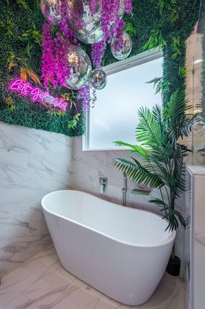 Modern luxe bathroom with freestanding white tub, marble tile, vertical green wall with purple blooms and mirrored disco balls, pink neon sign, and tropical plants by a frosted window
