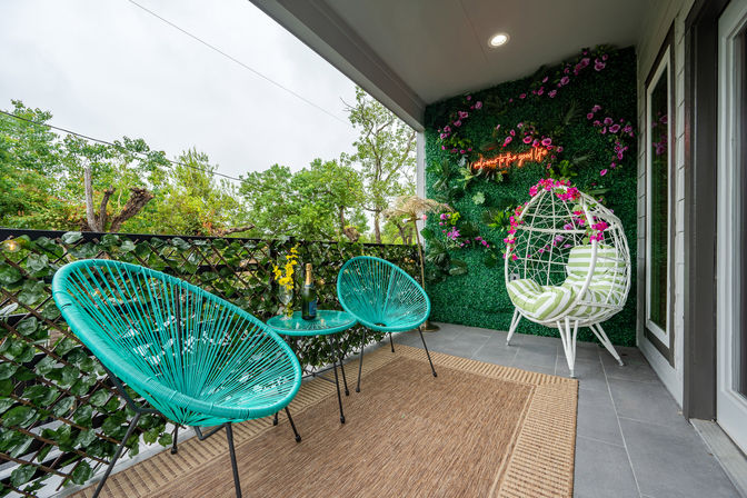 Cozy balcony patio with turquoise woven chairs, small table with bottle and glass, white hanging egg chair with striped cushion, lush green living wall with flowers and neon sign