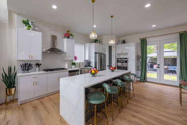 Bright modern kitchen with white marble waterfall island, gold-accent green bar stools, stainless steel appliances, pendant lights, wood floors, and French doors to a sunny backyard patio.