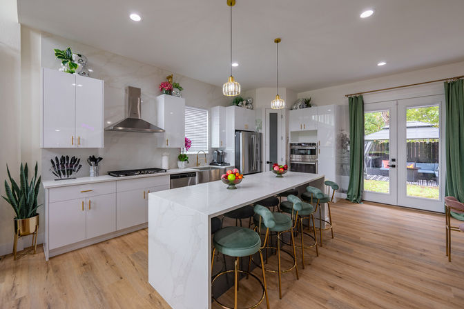 Bright modern kitchen with white marble waterfall island, gold-accent green bar stools, stainless steel appliances, pendant lights, wood floors, and French doors to a sunny backyard patio.