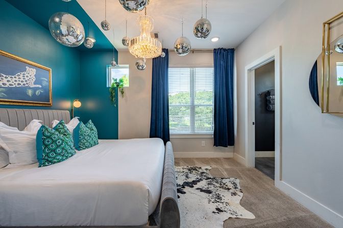 Bright modern bedroom with teal accent wall, upholstered bed with turquoise patterned pillows, crystal chandelier and hanging disco balls, large window with navy curtains and cowhide rug.