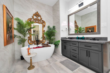 Spa-style luxury bathroom with a freestanding white tub filled with red rose petals, ornate gold baroque mirror, marble tile walls, tropical potted plants, small marble side table holding a bottle of rosé and glass, and a dark gray double-sink vanity with large framed mirror.