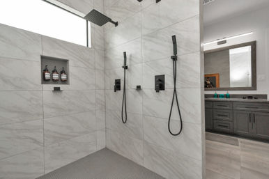 Contemporary marble-tiled walk-in shower with dual matte-black handheld showers and overhead rain head, recessed niche with amber shampoo bottles, and a gray vanity reflected in a mirror.