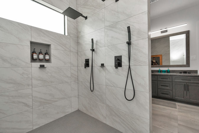 Contemporary marble-tiled walk-in shower with dual matte-black handheld showers and overhead rain head, recessed niche with amber shampoo bottles, and a gray vanity reflected in a mirror.