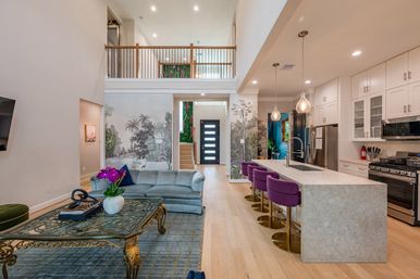 Bright modern open-concept living room and kitchen with two-story foyer, tropical mural accent wall, hardwood floors, marble island and purple bar stools