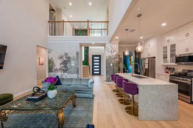 Bright modern open-concept living room and kitchen with two-story foyer, tropical mural accent wall, hardwood floors, marble island and purple bar stools