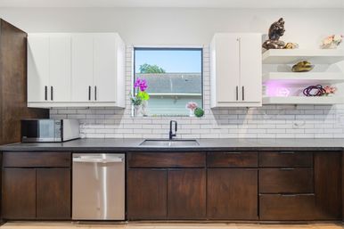 Bright modern home kitchen with white upper cabinets, dark wood lower cabinets, subway tile backsplash, stainless dishwasher, black faucet and a window sill with potted flowers.