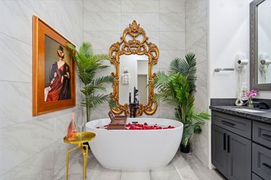 Luxury marble-tiled bathroom with a white freestanding tub sprinkled with red rose petals, ornate gold baroque mirror, tropical potted plants, a small gold side table with rosé and a glass, and dark gray vanity countertop.