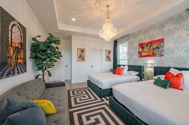 Boutique-style bright bedroom with two teal velvet platform beds, coral accent pillows, crystal chandelier, blue damask wallpaper, gray tufted sofa with yellow throw pillow, large potted plant and geometric rug