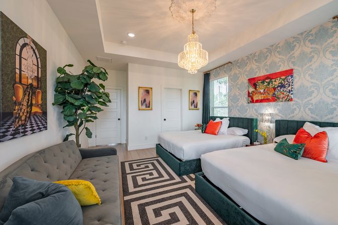 Boutique-style bright bedroom with two teal velvet platform beds, coral accent pillows, crystal chandelier, blue damask wallpaper, gray tufted sofa with yellow throw pillow, large potted plant and geometric rug