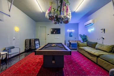 Vibrant game room with a black-felt pool table centered on a red patterned rug, green lounge sofa, foosball table, hanging disco balls with floral decor, neon wall signs and a wall-mounted TV