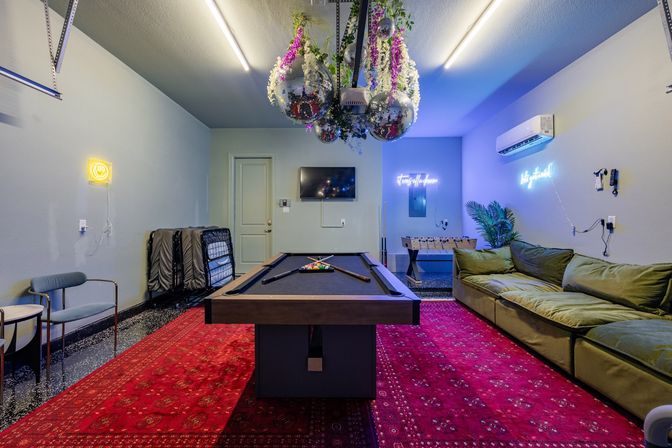 Vibrant game room with a black-felt pool table centered on a red patterned rug, green lounge sofa, foosball table, hanging disco balls with floral decor, neon wall signs and a wall-mounted TV