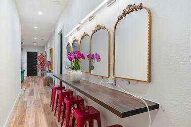 Bright residential hallway with white walls, long wooden counter beneath four ornate gold-framed mirrors and overhead lights, a vase of purple orchids, four red metal stools and warm hardwood flooring.