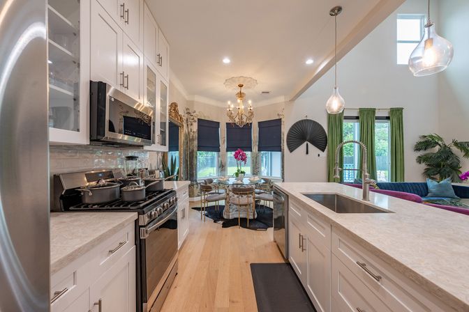 Open-concept modern kitchen in a suburban home with white shaker cabinets, stainless steel appliances, large island with marble-look countertop and spring faucet, pendant lights, hardwood floors and a bay dining nook with chandelier and navy Roman shades.