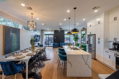 Contemporary open-concept kitchen and dining space featuring a large white marble island with teal barstools, black pendant lights, long dining table set for eight, hardwood floors, peacock wall art, white cabinetry and a bright living area with blue chairs and sliding glass doors.