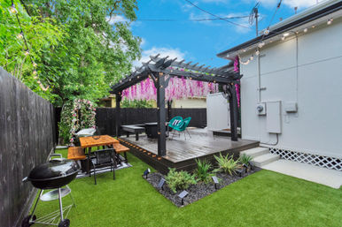 Cozy suburban backyard patio with a black pergola draped in pink hanging blossoms, string lights, wooden deck with turquoise chairs, picnic table, charcoal grill, and artificial turf