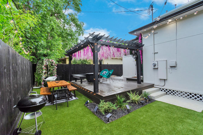 Cozy suburban backyard patio with a black pergola draped in pink hanging blossoms, string lights, wooden deck with turquoise chairs, picnic table, charcoal grill, and artificial turf