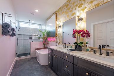 Modern tropical-inspired master bathroom with dark double-sink vanity and marble countertop, freestanding white tub, glass walk-in shower, gold palm wallpaper, pink orchid and neon accent light.