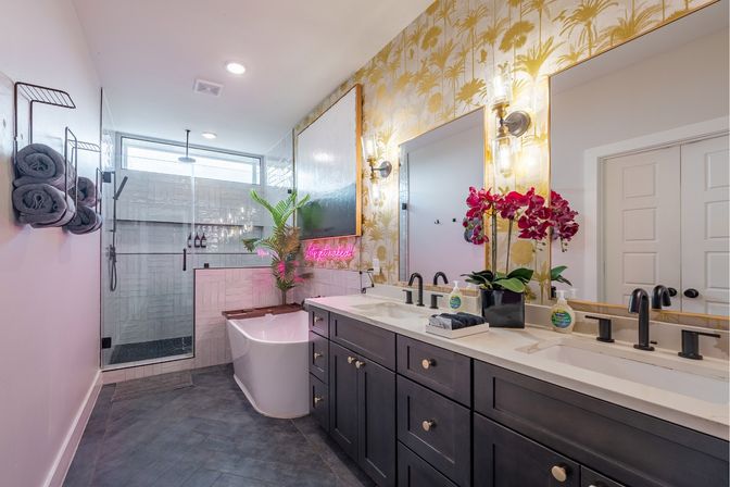Modern tropical-inspired master bathroom with dark double-sink vanity and marble countertop, freestanding white tub, glass walk-in shower, gold palm wallpaper, pink orchid and neon accent light.