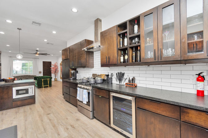 Bright open-concept modern kitchen with dark wood cabinets, white subway tile backsplash, stainless steel range and wine fridge, black countertops, light wood floors, island with built-in microwave, pendant lights, and a green velvet sofa visible in the adjoining living area.
