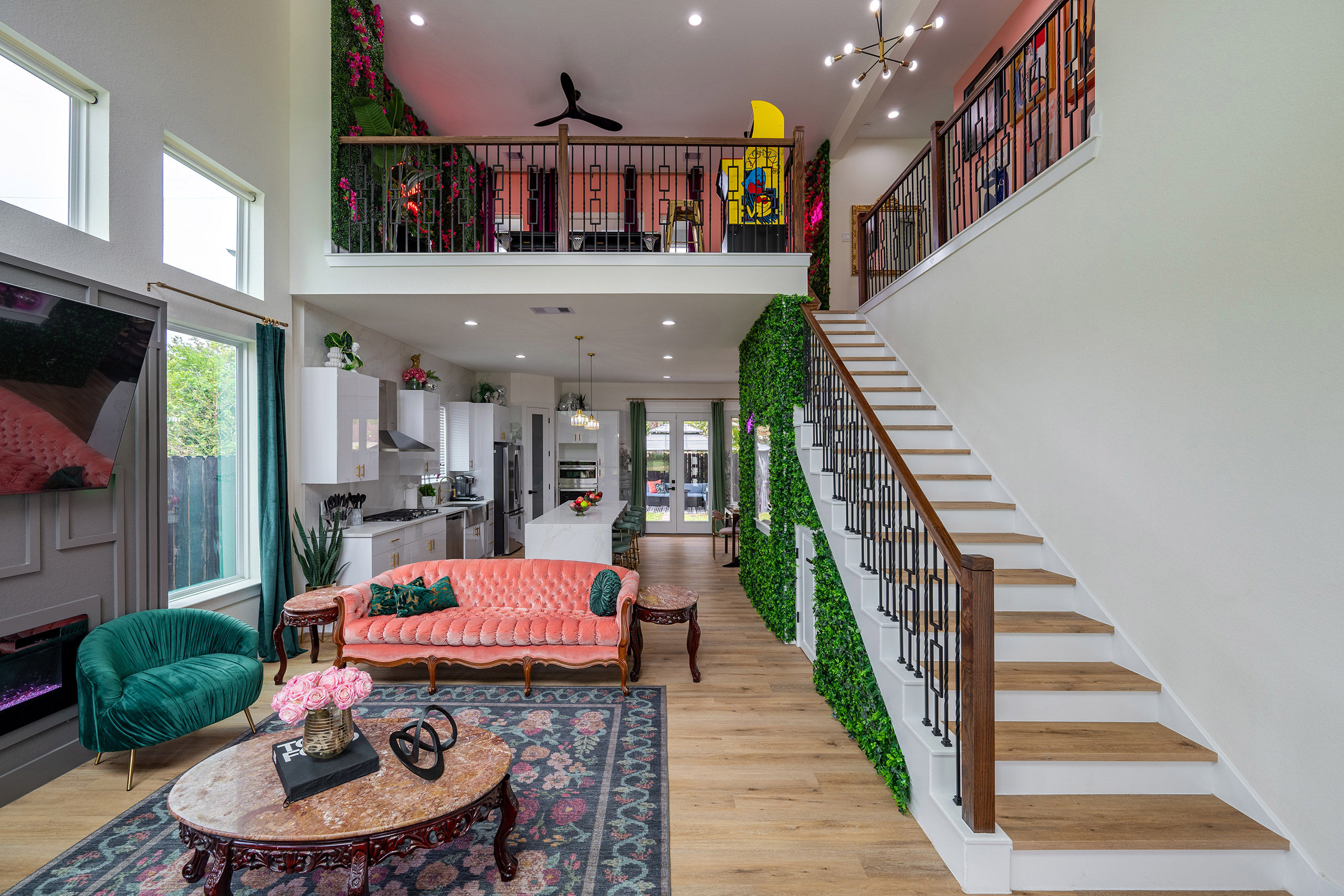 Modern open-concept two-story home interior with vibrant decor: coral tufted sofa, teal velvet chair, marble coffee table on a patterned rug, wood staircase with black metal balusters, green vertical plant wall, and a loft overlooking the white kitchen and island.