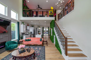 Modern open-concept two-story home interior with vibrant decor: coral tufted sofa, teal velvet chair, marble coffee table on a patterned rug, wood staircase with black metal balusters, green vertical plant wall, and a loft overlooking the white kitchen and island.