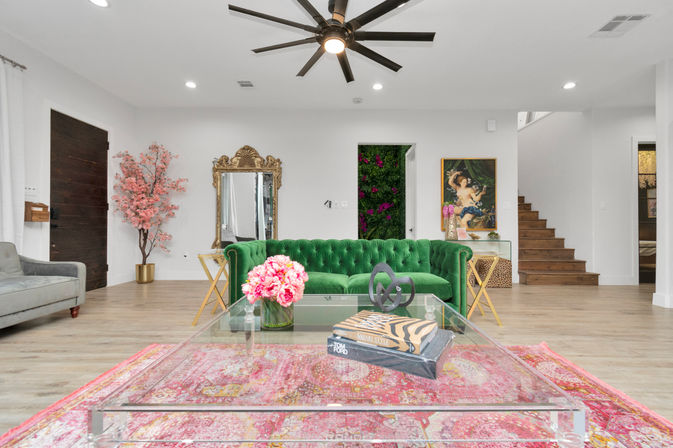 Bright modern open-concept living room with an emerald green tufted sofa, clear acrylic coffee table on a pink patterned rug, vase of pink flowers, ornate gold mirror, vertical floral accent, wooden staircase and large black ceiling fan.