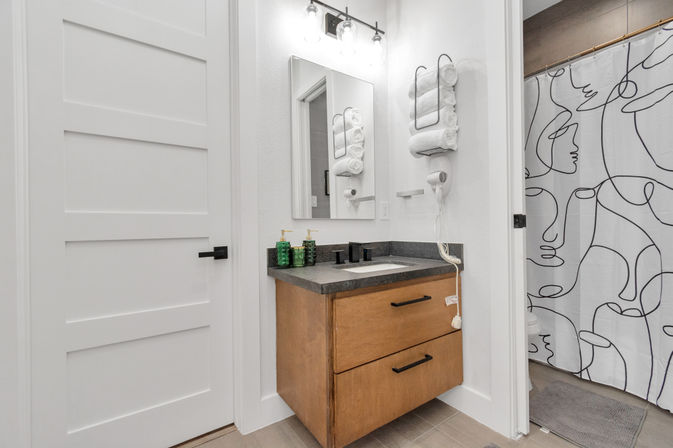 Bright contemporary bathroom with a floating wood vanity and black stone countertop, rectangular mirror and light fixture, wall-mounted rack of rolled white towels and hair dryer, green soap dispensers, and a shower curtain with abstract line-art faces.