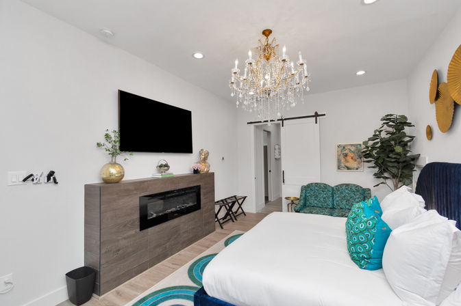 Bright modern bedroom with a sparkling crystal chandelier, wall-mounted TV above a wood-clad electric fireplace, white bed with teal peacock-print pillows, turquoise circular rug, green patterned loveseat, potted fiddle-leaf fig, and sliding barn door.