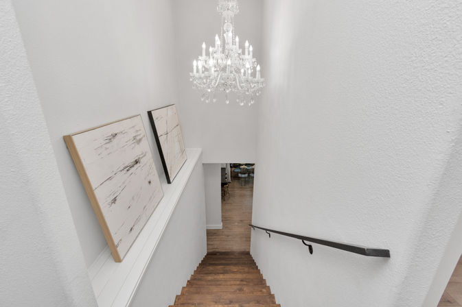 Interior view down a modern home staircase with dark wood steps, white textured walls, black metal handrail, sparkling crystal chandelier, and two leaning abstract canvases with an open dining area visible below.