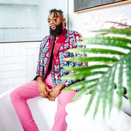 Person in a colorful teal-and-maroon patterned blazer and pink trousers seated in a bright white tiled bathroom with palm fronds in the foreground — playful fashion portrait.
