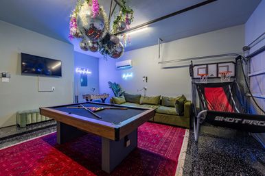 Playful modern game room with black-felt pool table, wall-mounted TV, foosball table, arcade basketball shooter, olive sectional on a red area rug, mirrored disco ball cluster and neon signs.