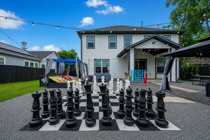 Backyard of a modern two-story home featuring a giant outdoor chess set with oversized black and white pieces on a checkerboard, a cabana lounge with colorful cushions, string lights overhead, grassy yard and covered patio under a bright blue sky
