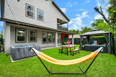 Suburban backyard of a modern two-story house with an orange hammock in the foreground, hot tub, wooden picnic table, covered gazebo lounge, string lights and fenced green lawn.