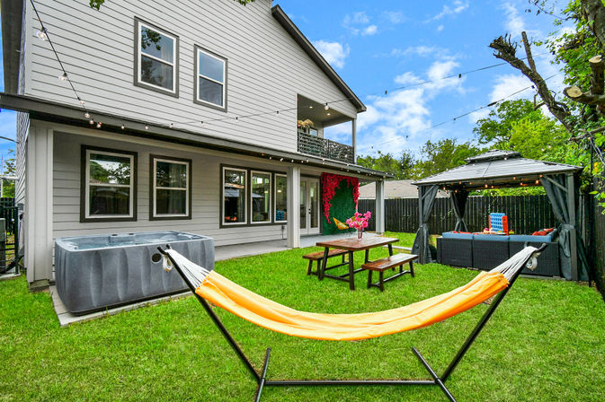Suburban backyard of a modern two-story house with an orange hammock in the foreground, hot tub, wooden picnic table, covered gazebo lounge, string lights and fenced green lawn.