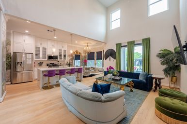 Bright open-concept living room and kitchen with double-height ceiling, white cabinetry and marble island with purple bar stools, velvet navy and light sofas, green curtains, gold-accent coffee table, light hardwood floors and sliding glass doors to a backyard.
