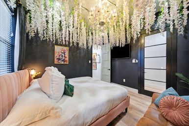 Stylish boutique apartment bedroom with pink velvet bed and white linens, cascading faux wisteria and crystal chandelier overhead, black accent wall with art, wall-mounted TV and leather sofa.