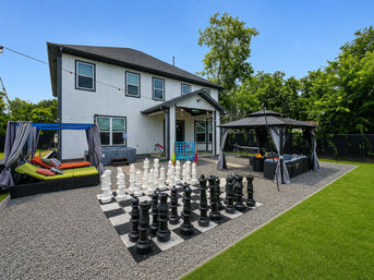 Modern two-story suburban backyard with oversized black-and-white chess set on a gravel patio, two curtained lounge cabanas with seating and a fire pit, hot tub, string lights, and well-kept green lawn