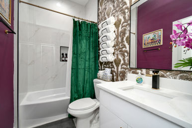 Small eclectic bathroom with shower-tub combo and green curtain, palm-print wallpaper with stacked rolled white towels, white vanity with black-and-gold faucet and pink orchid