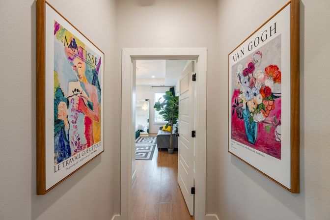 Narrow home hallway with colorful framed art prints on both walls, white door opening to a bright bedroom with hardwood floors, patterned rug, gray sofa and a tall potted plant